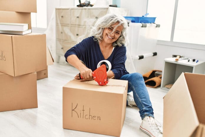 Middle age grey-haired woman smiling happy packing kitchen cardboard box at new home.