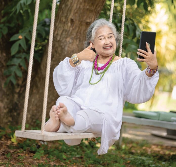 Elderly Woman Relaxing on a Swing While Using a Tablet in a Serene Outdoor Setting, Surrounded by Lush Greenery and Natural Beauty
