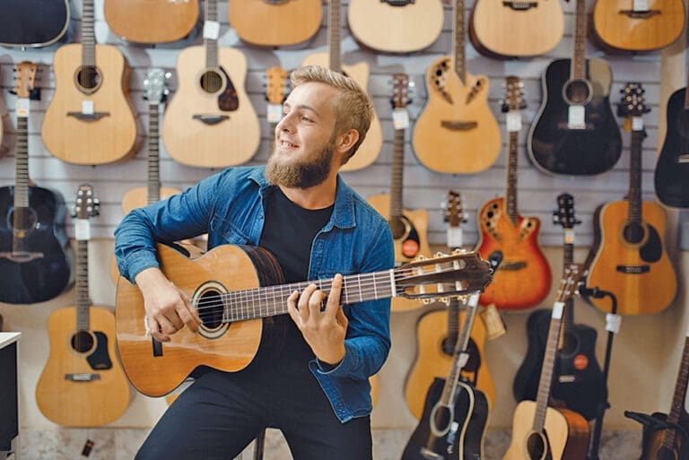 Young man plays on acoustic guitar in music store. Assortment in musical instruments shop, male musician buying equipment