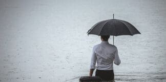Rear view on a handsome man wearing white shirt and holding umbrella and a briefcase, standing in the water on a rainy day. The man waded knee-deep in water. Selective focus, three quarter length, copy space has been left.