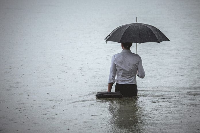 Handsome man standing in water and holding umbrella during rain Rear view on a handsome man wearing white shirt and holding umbrella and a briefcase, standing in the water on a rainy day. The man waded knee-deep in water. Selective focus, three quarter length, copy space has been left.