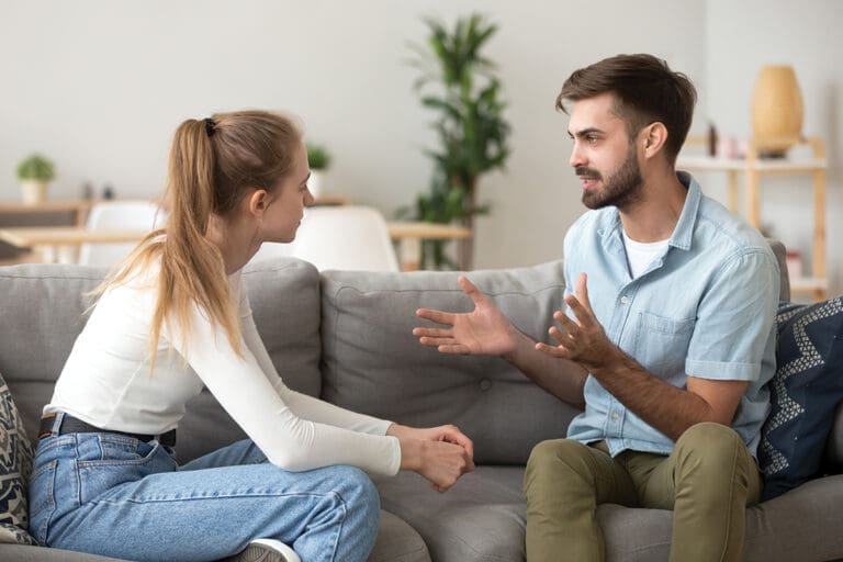 Serious young couple sitting together on sofa, talking about relationships, spending time together at home, focused wife listening to speaking husband, friends having conversation